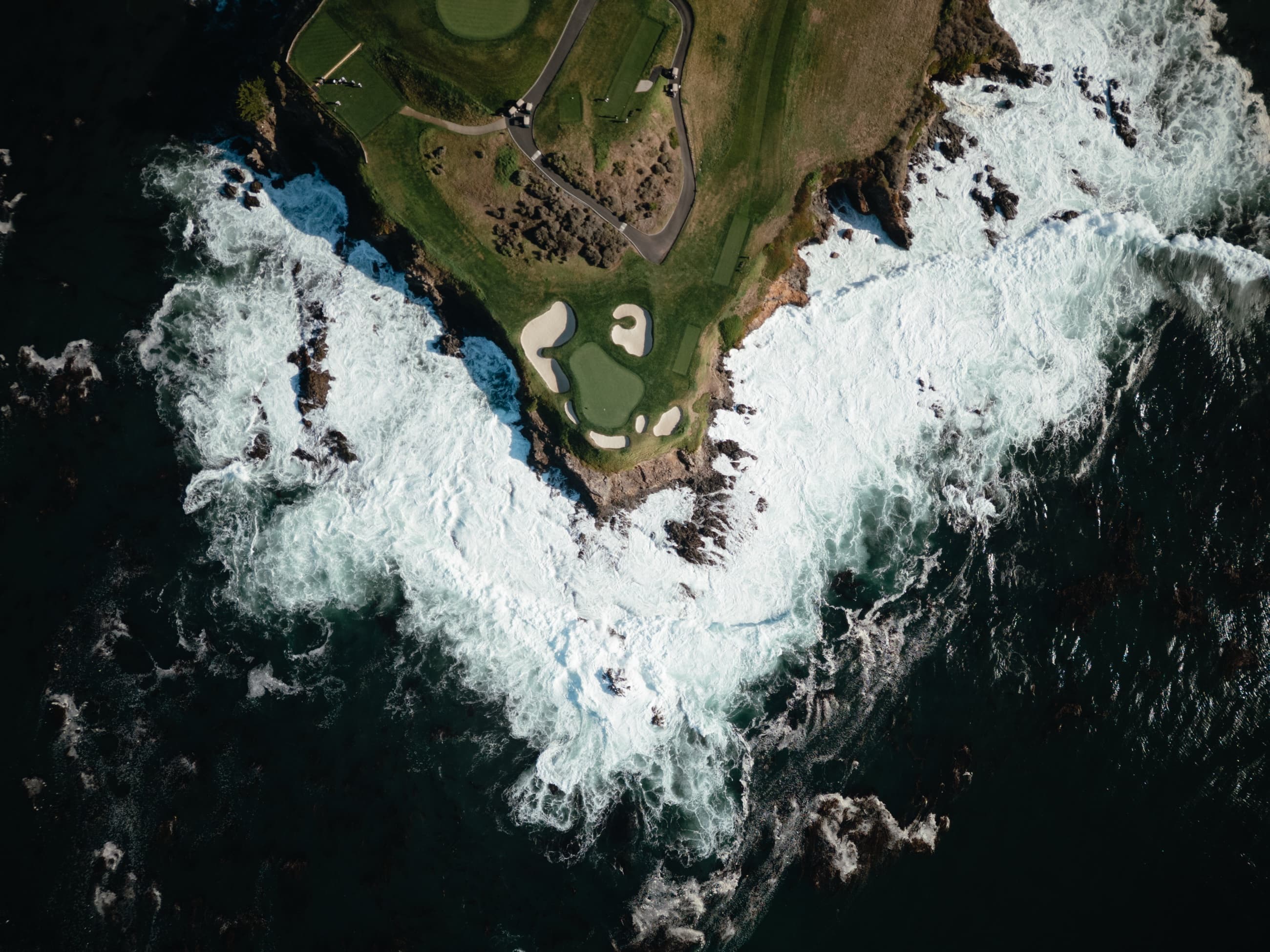 A cliffside golf green ringed by crashing Pacific surf at Pebble Beach, from above