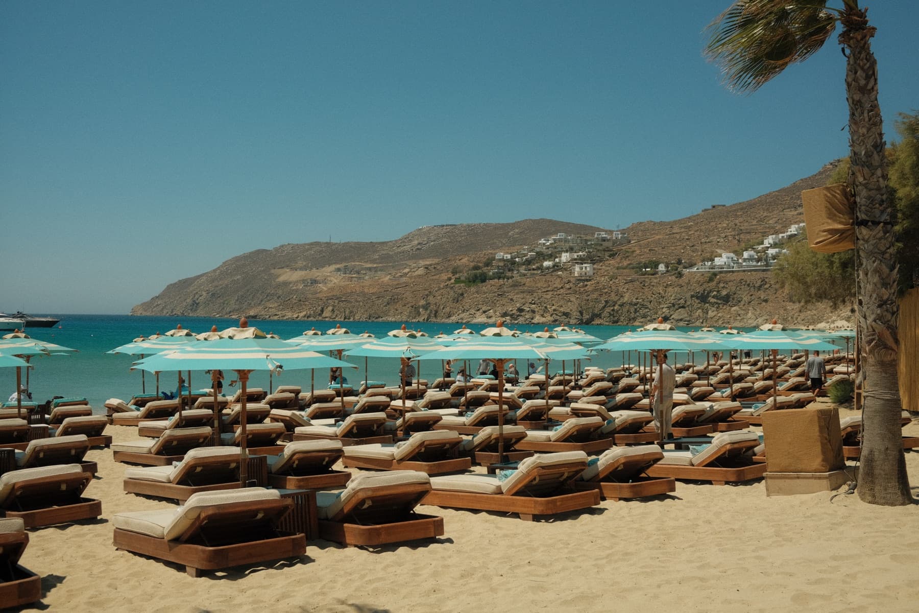 Rows of empty sun loungers under soft blue umbrellas on a quiet Greek beach, the sea and hills beyond